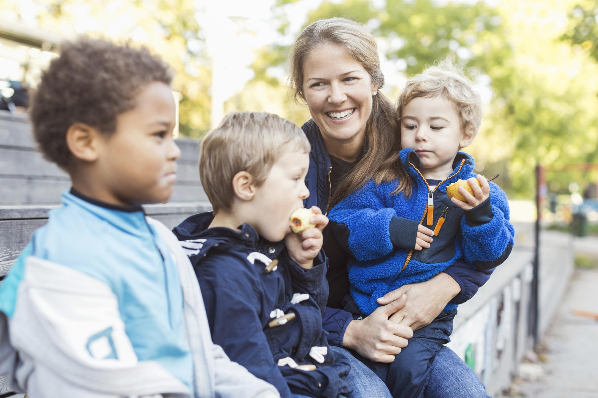 Tre børn sidder udenfor og spiser frugt med en smilende voksen 