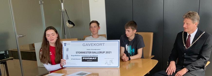 Tre børn sidder bag ved en stor check. Borgmesteren sidder ved siden af