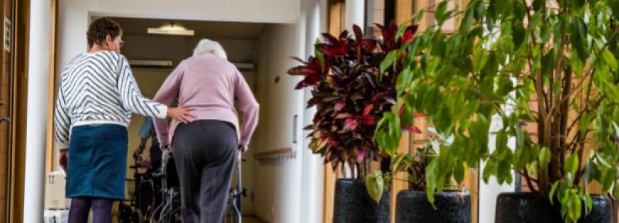 Ældre dame med rollator får støtte, mens hun går væk fra kameraet